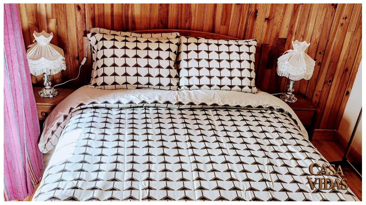 Cama com colcha e fronhas geométricas pretas e brancas, em quarto com madeira rústica e abajures.